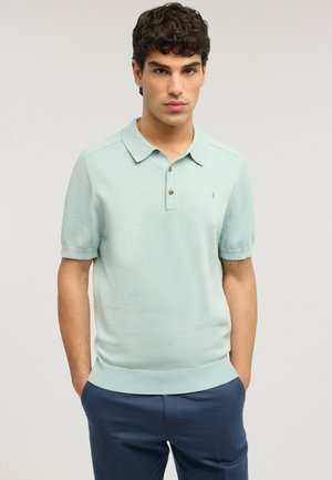 Młody mężczyzna o krótkich, kręconych włosach, ubrany w jasnooliwkową koszulkę polo z krótkim rękawem i ciemnoniebieskie spodnie, z rękami w kieszeniach, na neutralnym tle.