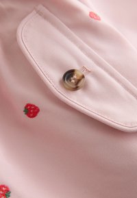 Närbild av en rosa tygficka med en brun knapp och små röda jordgubbsmönster broderade på tyget.