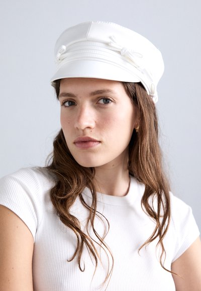 Weißer Hut aus glattem Material mit dekorativen Bändern und strukturierten Streifen. Das Modell trägt ein geripptes weißes Shirt und hat lange, wellige Haare.