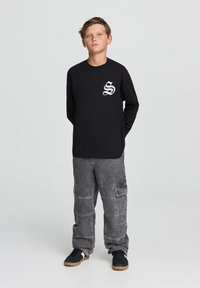 Czarna koszula z długim rękawem z białym logo graficznym na piersi, zestawiona z szarymi spodniami cargo z wieloma kieszeniami i luźnym krojem.