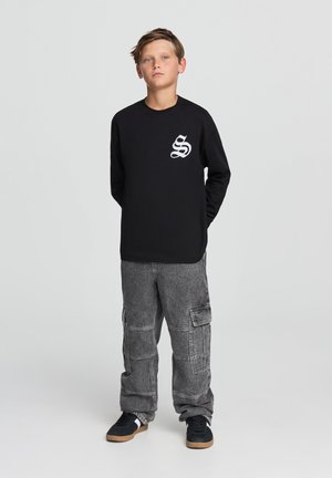Maglietta nera a maniche lunghe con logo grafico bianco sul petto, abbinata a pantaloni cargo grigi con molte tasche e una vestibilità ampia.