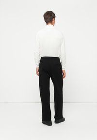 Weißes Langarmoberteil und schwarze Hose mit elastischem Bund, mit einer Gesäßtasche. Das Model wird von hinten vor einem schlichten Hintergrund gezeigt.