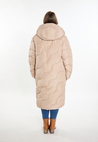 Cappotto lungo e imbottito di un beige chiaro con un design a trapunte. Caratterizzato da un cappuccio e maniche oversize, abbinato a jeans blu e stivali con tacco marroni.