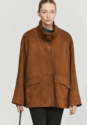 Frau trägt eine lockere braune Velourslederjacke mit hohem Kragen und großen Fronttaschen, hält einen schwarzen Handtaschenriemen in der linken Hand.