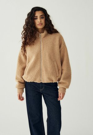 Kobieta z długimi kręconymi włosami ubrana w beżową polarową kurtkę na zamek i ciemnoniebieskie dżinsy, stojąca na tle jednolitego jasnego tła.