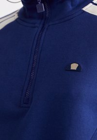 Maglione blu navy con zip, caratterizzato da un colletto alto, polsini elasticizzati e un piccolo logo ricamato in crema e blu scuro sul petto.