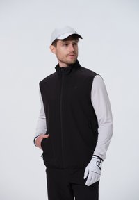 Zwarte mouwloze vest met een hoge kraag, gecombineerd met een wit overhemd met lange mouwen. Het model draagt een lichtgrijze pet en witte golfhandschoenen.