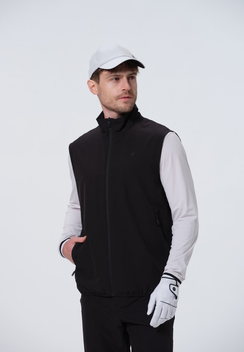 Zwarte mouwloze vest met een hoge kraag, gecombineerd met een wit overhemd met lange mouwen. Het model draagt een lichtgrijze pet en witte golfhandschoenen.