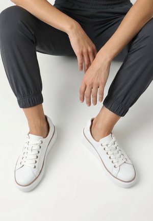 Personne assise, jambes écartées, portant un jogging noir et des baskets blanches à lacets sur un fond de sol blanc.