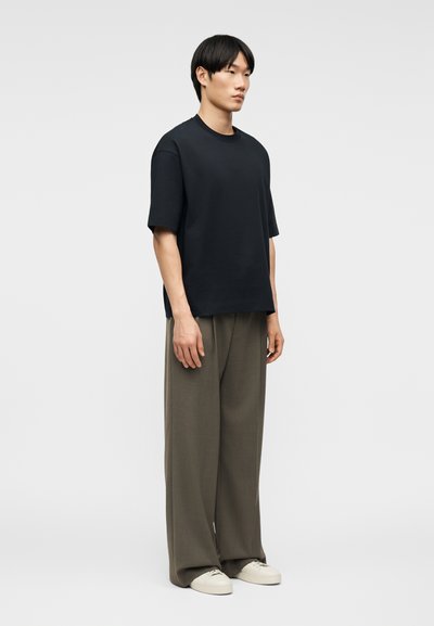 Νεαρός άνδρας που στέκεται, φορώντας ένα φαρδύ μαύρο T-shirt, φαρδιά χακί παντελόνια και λευκά αθλητικά παπούτσια μπροστά σε ένα απλό ανοιχτό φόντο.