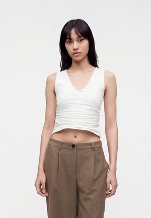Femme aux longs cheveux foncés portant un top court blanc sans manches et un pantalon marron taille haute, debout devant un fond uni.
