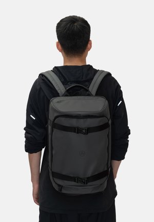 Zaino grigio realizzato in nylon resistente, con forma rettangolare, spallacci regolabili e accenti neri con un dettaglio logo centrale.