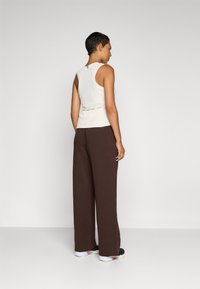 Light beige sleeveless top with a fitted waist and subtle red stitching, paired with loose brown trousers and black sneakers.