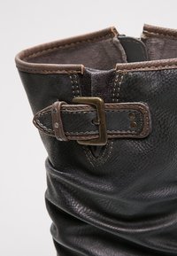 Stivaletto in pelle nera con design scivolato, caratterizzato da una fibbia e una cinghia marroni, cuciture visibili e una superficie texturizzata.