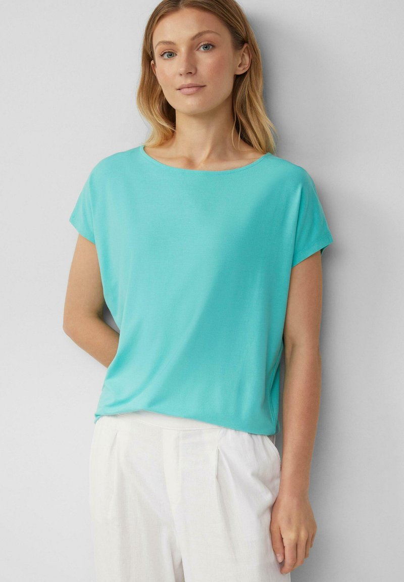 Vrouw in een licht turquoise top met korte mouwen en witte high-waisted broeken, staand tegen een effen lichtgrijze achtergrond.