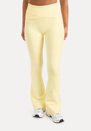 Person iført blege gule højtaljede flare leggings og hvide sneakers, stående mod en ensformig lys baggrund.