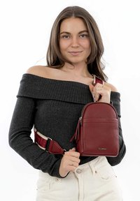 Červená kožená křížová taška s texturovaným povrchem, zipovým uzávěrem a nastavitelným popruhem. Modelka má na sobě šedý svetr s odhalenými rameny a světlé kalhoty.