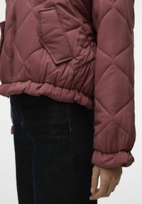 Giacca bomber trapuntata bordeaux con tasca laterale e polsini elastici; presenta un orlo a costine e una texture liscia e lucida.