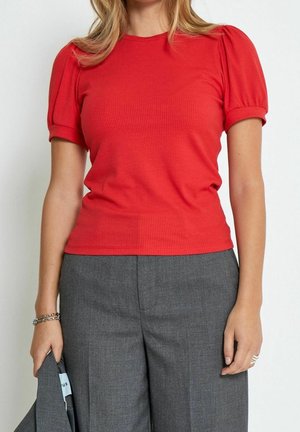 Femme portant un haut rouge à manches courtes côtelé avec des manches bouffantes et un pantalon gris taille haute, tenant une veste grise assortie.