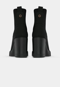 Crne čizme do članaka na petu imaju glatki materijal, rebrasti gornji dio sličan čarapi i teksturiranu petu. Ukrasni detalji uključuju okrugli logo.