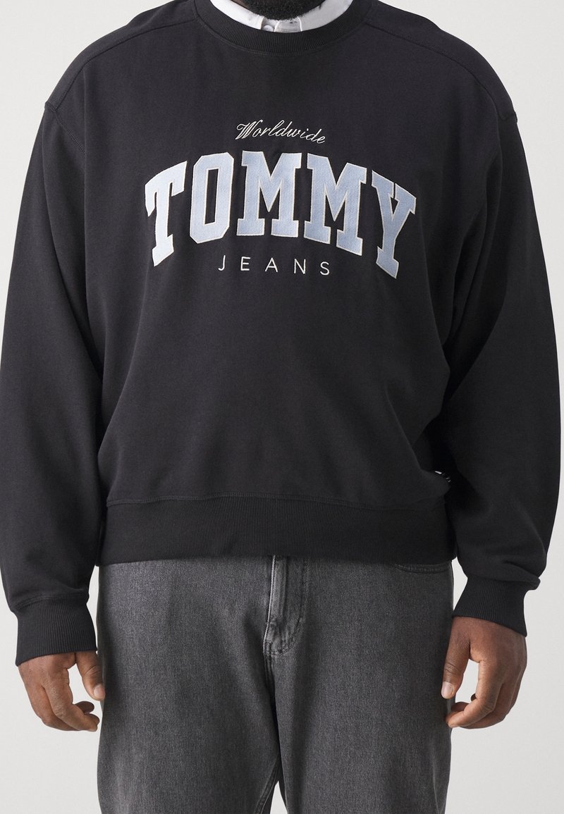 Personne portant un sweat-shirt noir avec un grand texte "TOMMY" et un texte plus petit "Worldwide Jeans", associé à un jean en denim gris.