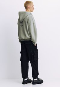 Hanorac verde supradimensionat cu un logo pe spate, asortat cu pantaloni cargo negri cu mai multe buzunare și manșete elastice, purtat cu pantofi negri.