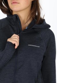 Navy zip-up hoodie with textured fabric, a fitted design, and a logo on the chest. Features a hood and high collar.