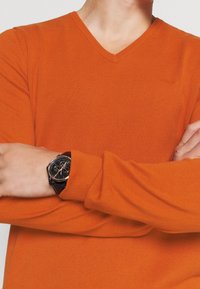 Oranžový svetr s výstřihem do V vyrobený z texturované pleteniny. Na levém zápěstí je vidět černé hodinky s kulatým ciferníkem a stř�íbrnými akcenty.