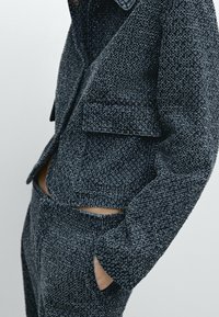 Giacca scura intrecciata con tessuto testurizzato, caratterizzata da due tasche frontali e design corto. Abbinata a pantaloni coordinati, entrambi di colore simile.