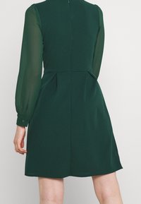 Grön klänning i slätt tyg, med långa genomskinliga ärmar, hög halsringning och plisserade detaljer på baksidan, som når till mitten av låret.