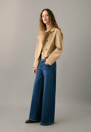 Mujer con cabello largo y ondulado que lleva una chaqueta beige corta, camiseta blanca, vaqueros azules de pierna ancha y zapatos negros, de pie contra un fondo liso.