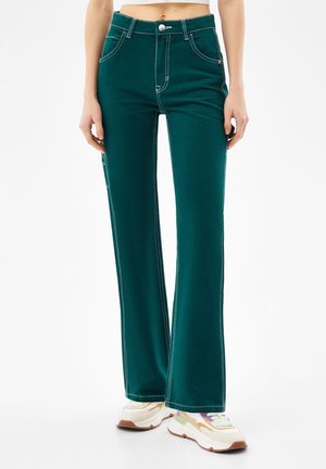 Frau trägt dunkelgrüne High-Waist-Jeans mit kontrastierenden weißen Nähten und weißen klobigen Sneakers, steht vor weißem Hintergrund.