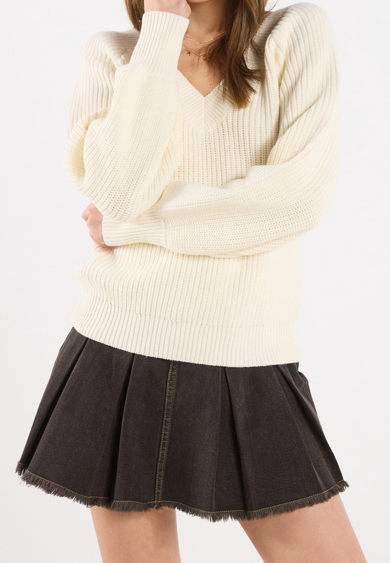 Pull tricoté couleur crème avec un col en V et un ourlet côtelé, associé à une jupe plissée en denim noir avec un bord à franges.