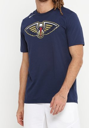 Mann trägt ein marineblaues Nike-T-Shirt mit einem großen Logo des Basketballteams New Orleans Pelicans und weißen Shorts.