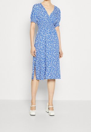 Femme portant une robe midi bleue à imprimé floral blanc, manches courtes bouffantes, une fente latérale et des chaussures à plateforme Mary Jane blanches.