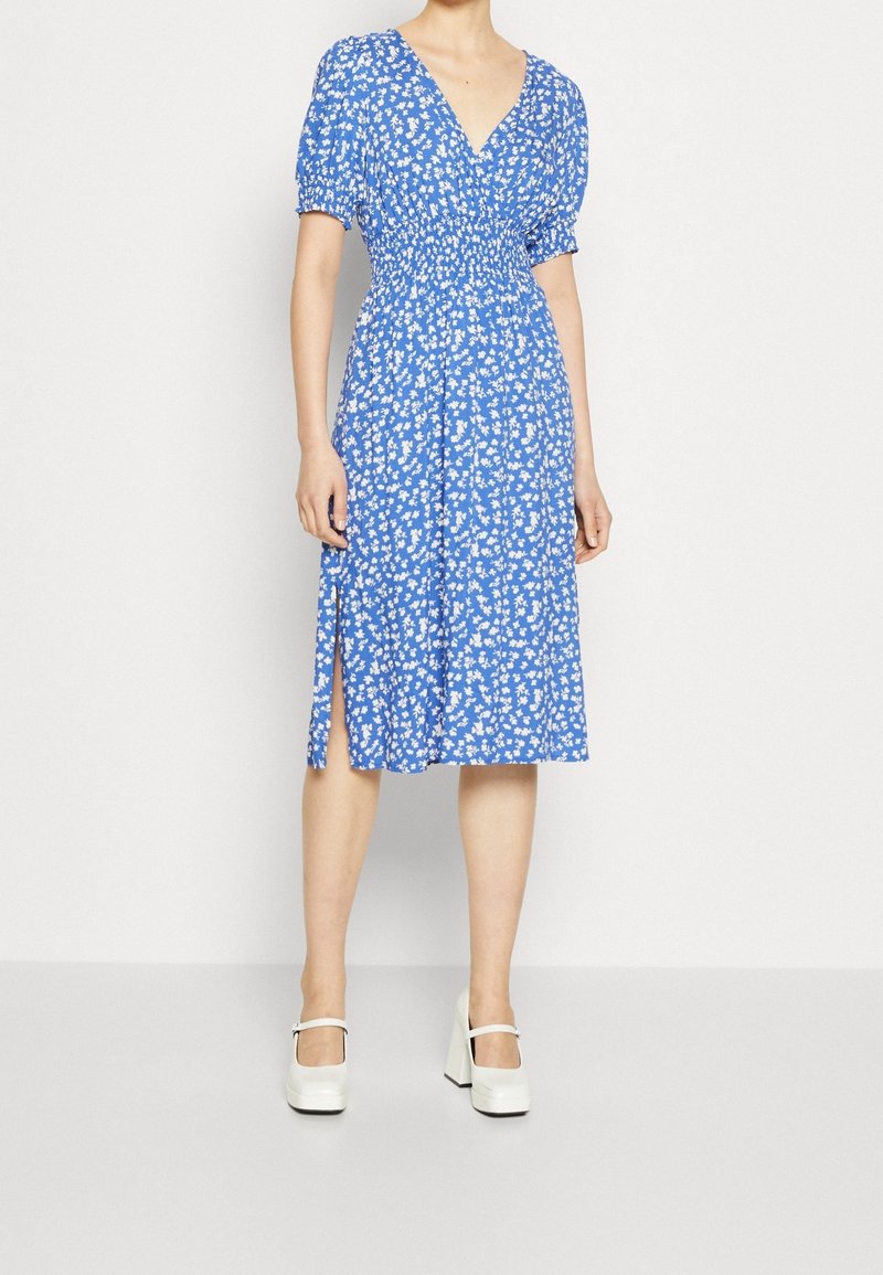 Femme portant une robe midi bleue à imprimé floral blanc, manches courtes bouffantes, une fente latérale et des chaussures à plateforme Mary Jane blanches.