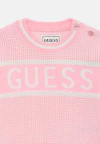 Rosa strikkegenser med hvite striper og "GUESS"-logo. Har en ribbestrikket krage og to knapper på skulderen for lukking.