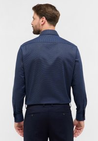 Uomo con capelli castani e barba visto da dietro, che indossa una camicia strutturata blu scuro e pantaloni blu scuro su uno sfondo neutro.