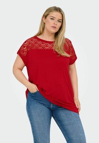 Rød top lavet af blødt stof, med en tekstureret blonder-yoke i et geometrisk mønster. Kortærmede og afslappet fit, parret med blå jeans.