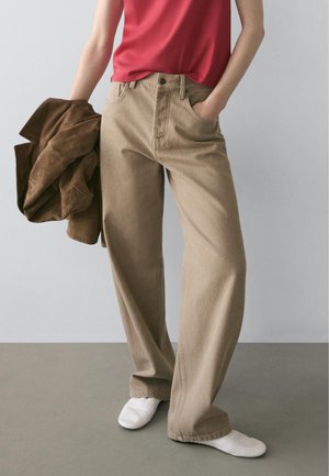 Person trägt beige weite Hose, rotes ärmelloses Oberteil, weiße Schuhe, hält eine braune Jacke in der linken Hand vor einem schlichten Hintergrund.