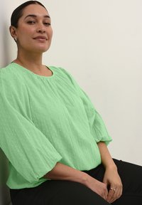 Mentolově zelená blůza s texturovanými tečkami, kulatým výstřihem a volnými rukávy, spárovaná s černými kalhotami. Model sedí proti světlé zdi.
