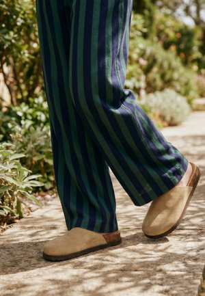 Personne portant un pantalon rayé bleu marine et vert marchant sur un chemin en pierre avec des chaussures beige sans lacets et de la verdure en arrière-plan.