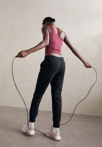 Persoon in een roze tanktop en zwarte broek die binnen springt met een springtouw op een grijze vloer tegen een beige muur.