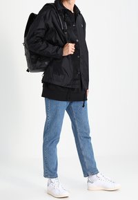 Svart lättviktsjacka med tryckknappar, kombinerad med en svart sweatshirt, blå jeans och vita sneakers. Svart ryggsäck med remmar.