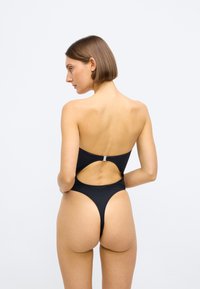 Μαύρο ολόσωμο μαγιό με άνοιγμα στην πλάτη και ασημί λεπτομέρειες. Λείο ύφασμα και μινιμαλιστικός σχεδιασμός. Υψηλές σχισμές στα πόδια.