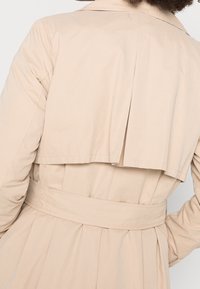 Béžový trench coat s vysokým límcem, dlouhými rukávy a páskem v pase. Vyznačuje se vrstveným designem zadní části s skladkami. Hladká, lehká látka.