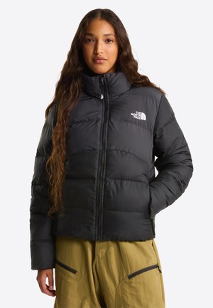 Kobieta w czarnej puchowej kurtce North Face z zamykanymi kieszeniami i oliwkowozielonych spodniach, stojąca z jedną ręką w kieszeni.