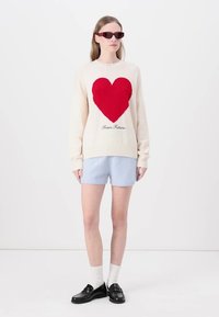 Kremgenser med et rødt strikket hjerte-design og teksten "Fra Fremtiden", kombinert med lyseblå shortser og svarte loafers.