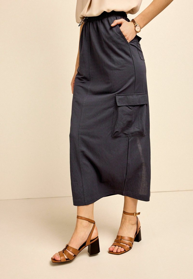 Jupe midi bleu marine avec taille élastique, poches latérales et une poche plaquée sur la gauche. Sandales à talons carrés marron complètent le look.