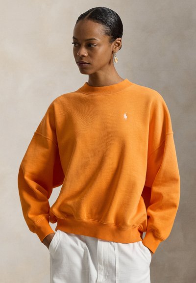 Πορτοκαλί βαμβακερό φούτερ με ριμπ λαιμόκοψη και μανίκια με μανσέτες. Διαθέτει μικρό λευκό λογότυπο στο στήθος. Φοριέται με λευκό παντελόνι.